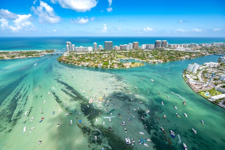 Aerial view of a coastal cityscape with boats in turquoise water and skyline in the background.