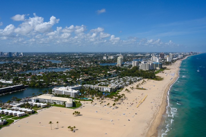 Aerial view of a long sandy beach with nearby cityscape and ocean under a clear blue sky.