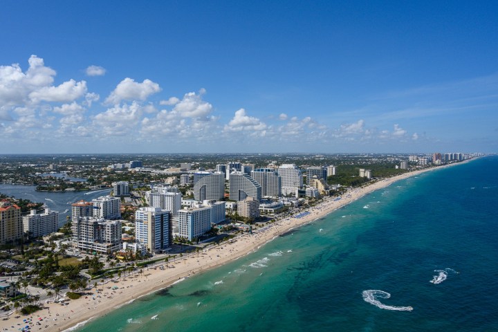 Aerial view of a city skyline along a beach with turquoise ocean waves under a blue sky with scattered clouds.