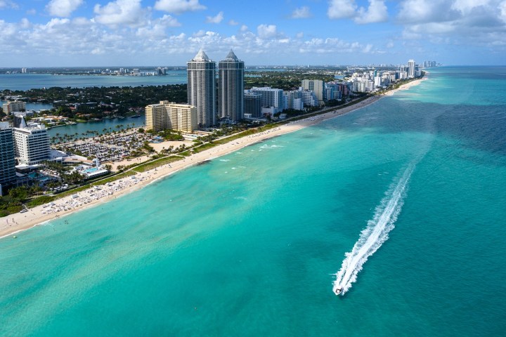 Aerial view of a city skyline along a beach with turquoise water and a speeding boat.