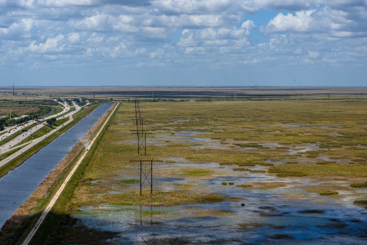 Highway beside a canal with power lines and wetlands under a cloudy sky.