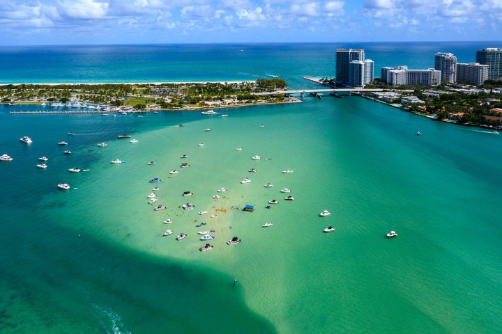 Aerial view of boats gathered in turquoise water near an urban shoreline with buildings and a bridge.