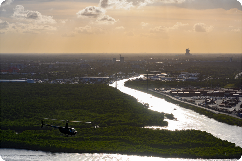 Helicopter flying over river and greenery, city skyline in the background at sunset.