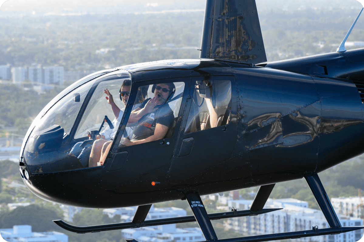 Two people inside a flying helicopter, with one waving and wearing sunglasses.