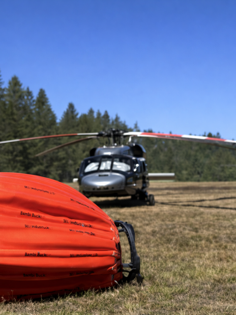 Helicopter on grass field with orange firefighting bucket in foreground.