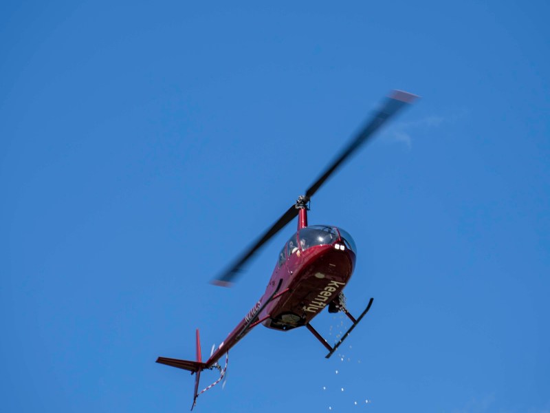 Red helicopter flying against blue sky, releasing small particles downward.