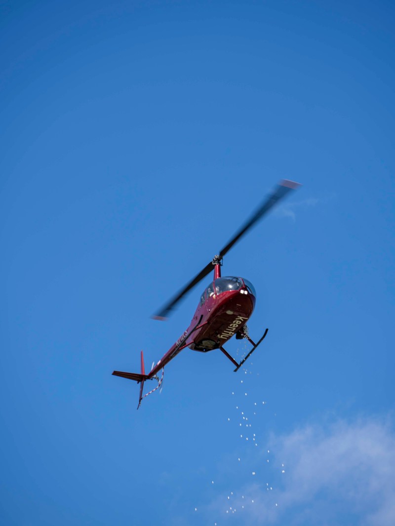 Red helicopter flying against blue sky, releasing small particles downward.