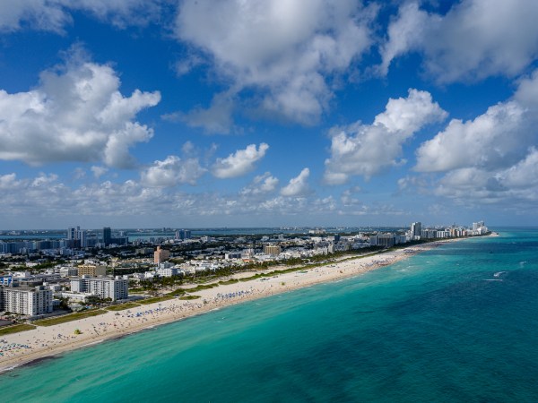 Aerial view of a beach with turquoise water, city skyline, and scattered clouds.