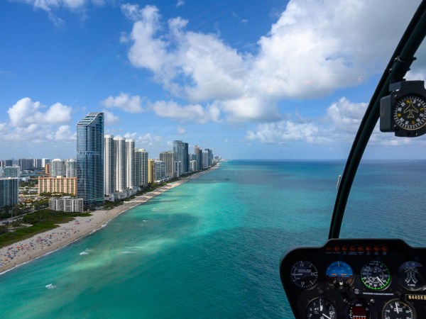 Aerial view of a city coastline with skyscrapers and turquoise ocean, seen from a helicopter cockpit.