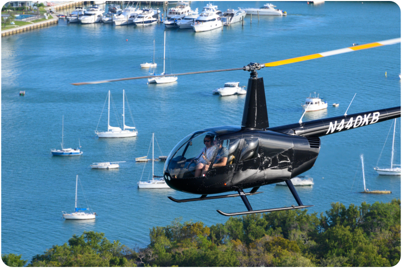 Black helicopter flying over a marina with several yachts and sailboats on blue water.