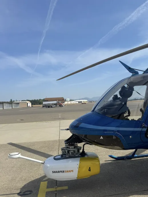 Blue helicopter parked on tarmac with clear skies and runway in background.