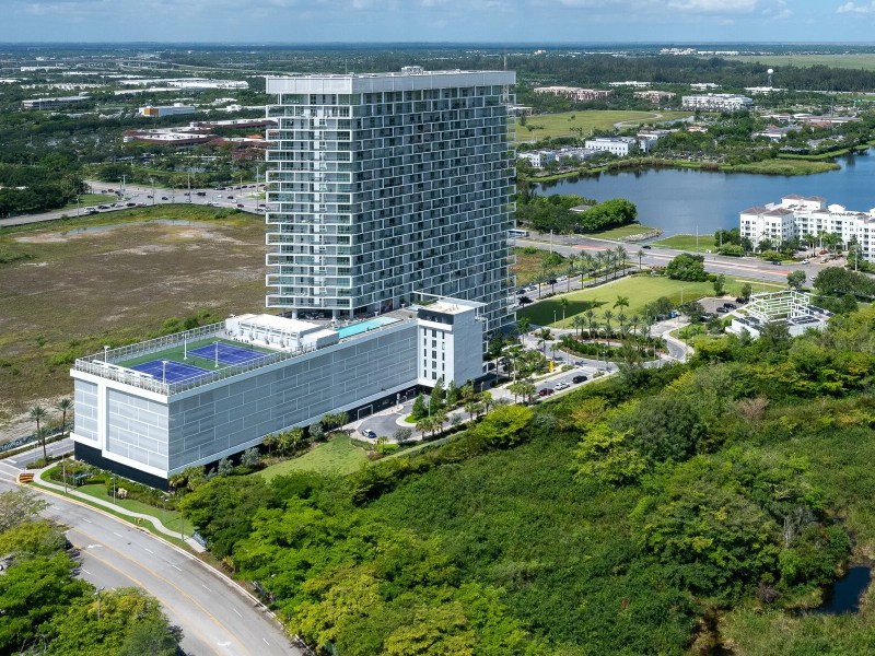 Aerial view of a tall modern building with a rooftop pool, surrounded by greenery and water.