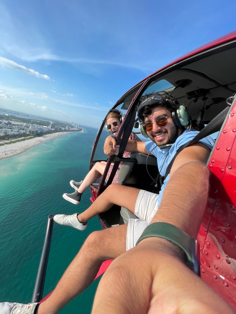 Two people in a helicopter flying over a coastline, smiling and taking a selfie.
