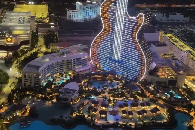 Guitar-shaped building lit up at night, surrounded by palm trees and a lake.